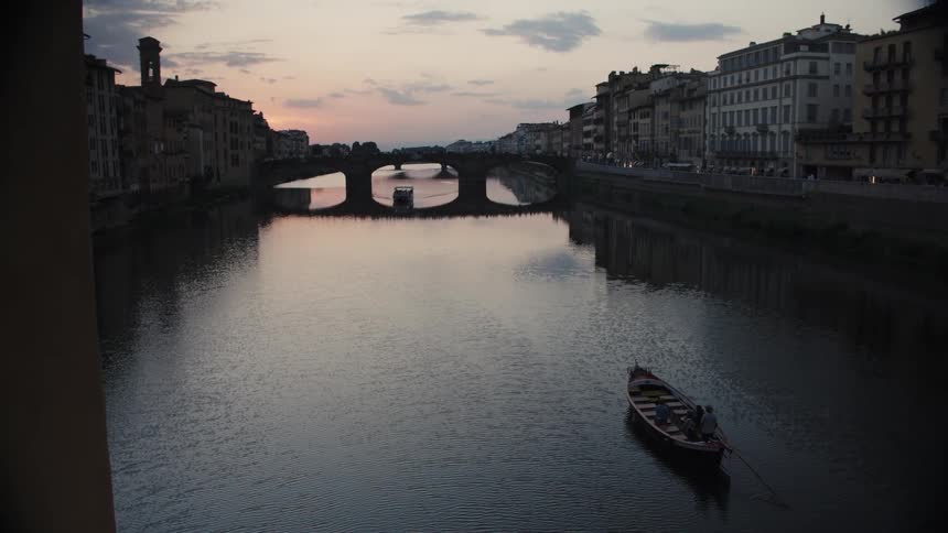 Boat sails on a calm river at sunset with a bridge and historic buildings in the background