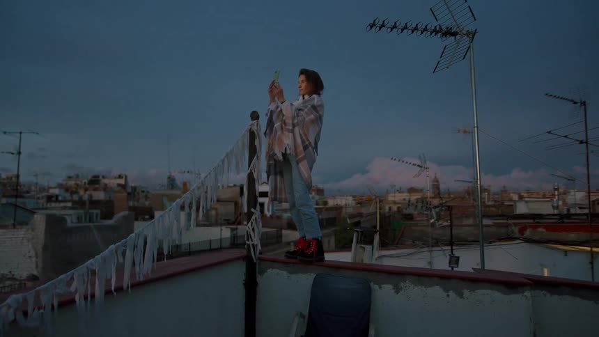 A young woman in jeans and a shawl stands on a rooftop ledge against a dusk sky holding a smartphone to take a picture of the cityscape below which is covered in antennae and rooftops with laundry or fabric hanging
