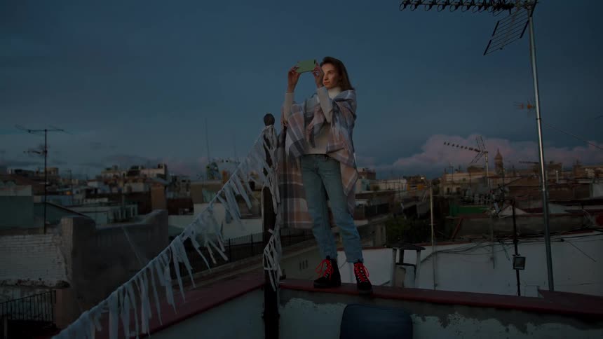 Woman on a ledge at dusk photographing the city with her phone