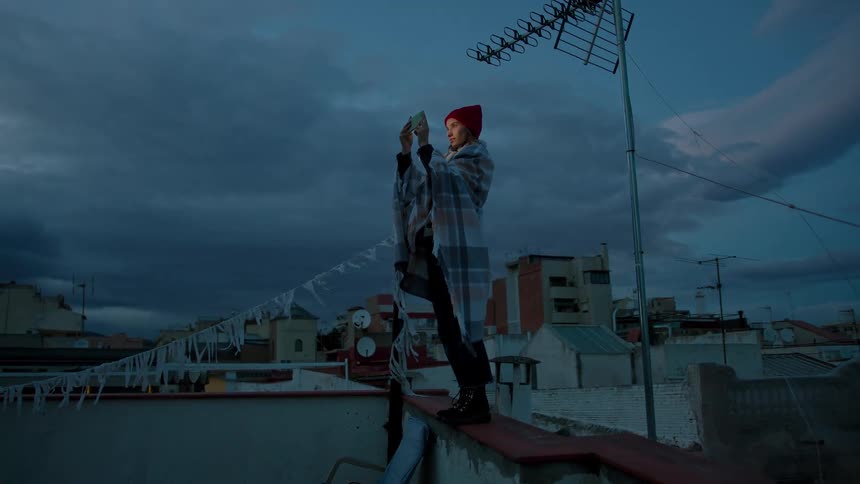Woman wearing a red beanie and blanket takes a photo with her phone on a rooftop