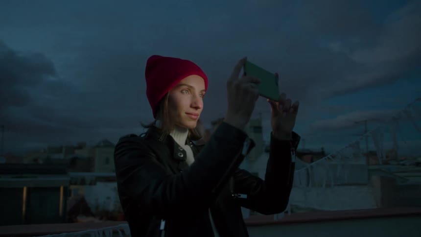 A woman in a red beanie captures the twilight city skyline with her smartphone from a rooftop