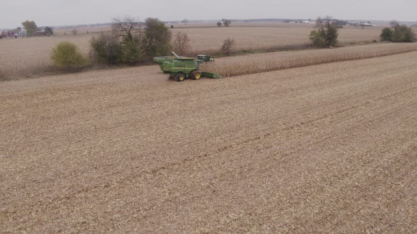 A combine harvester and a tractor with a grain cart working together to harvest a vast dry cornfield under an overcast sky
