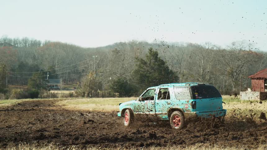 A teal suv fishtails through a muddy field flinging clumps of dirt into the air with bare trees and low hills in the background
