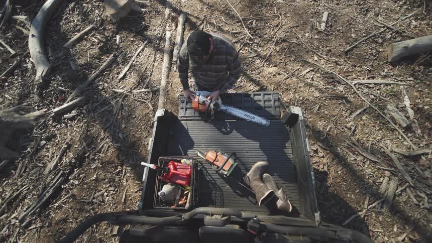 A young man walks up to the rear cargo bed of a utility vehicle leans over the tailgate and lifts a chainsaw that is stored next to a fuel container and a pair of boots