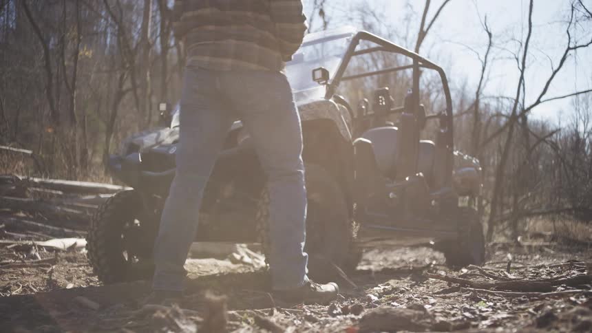 A bearded man in a plaid jacket walks near an utv in a leafless forest looking focused as he grabs his hat and gets inside the utv