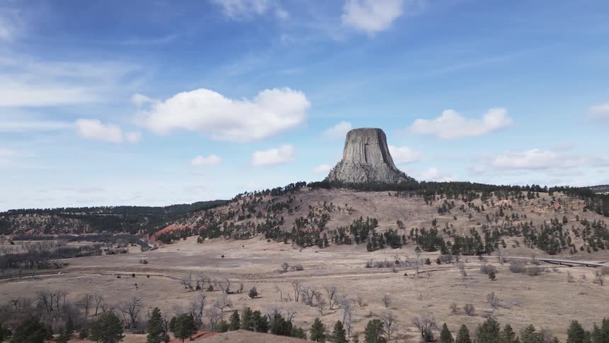 An aerial view video shows the enormous dark towering rock formation of devils tower rising above a sprawling landscape of brown sparsely forested hills and flatlands under a bright blue sky with moving white clouds