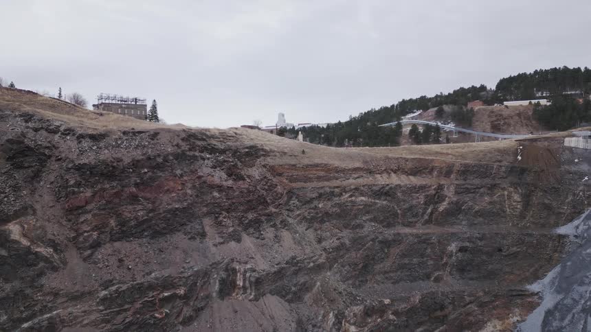An aerial view moves over the rocky terraced wall of a massive openpit mine revealing a steep vertical structure of bluegrey material on the right and then pans to show scattered buildings and a slide structure on the hillside above the excavation