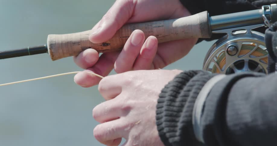 Closeup of hands working a flyfishing rod and reel yellow line stretched tight beside blue water