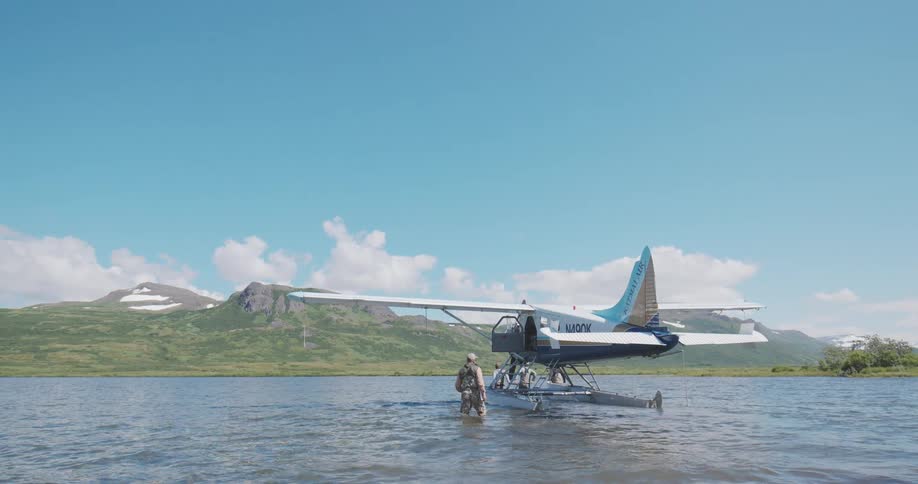 In a breathtaking mountain scene a person in waders approaches a floatplane on a tranquil lake