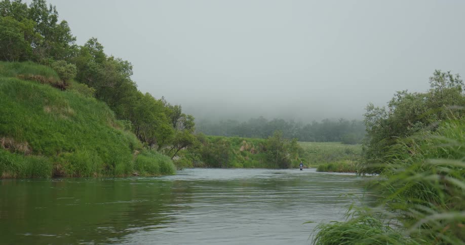 The video shows a wide static shot of a river flowing through a lush green valley on what appears to be a foggy or overcast daythe river is flanked by a steep grassy bank on the left and dense foliage on the right with a small blue figure likely