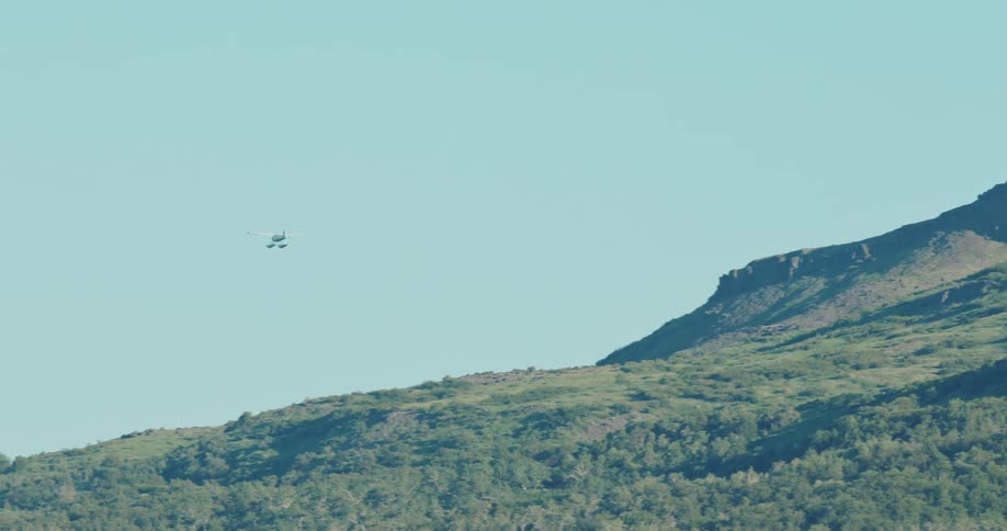 A small aircraft flies horizontally in a light blue sky past a large steep mountain ridge covered in green scrub