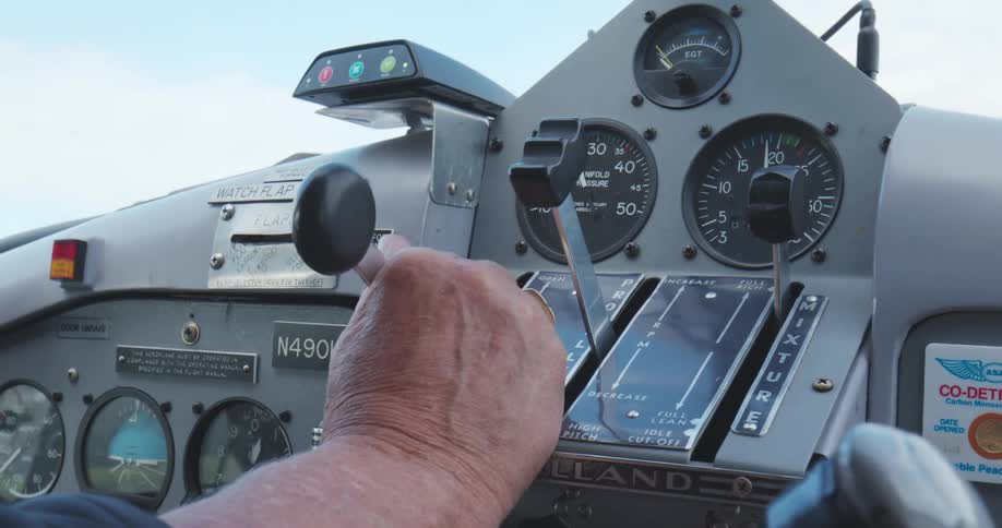 A human hand with visible veins moves a lever on the control panel of a small aircraft cockpit that contains multiple gauges and instrument labels