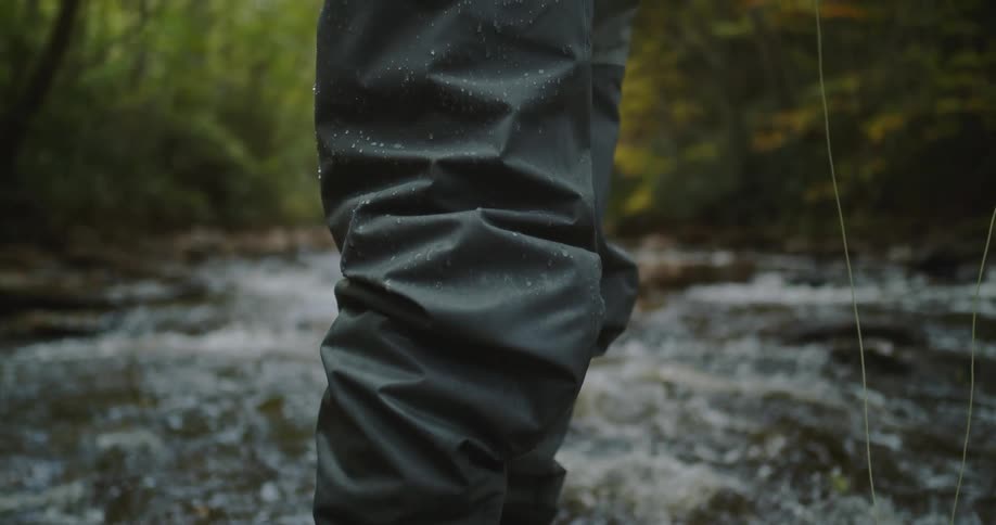A person wearing dark waders and fishing boots stands on a wet rock in the middle of a flowing river retrieves a fishing line and holds a circular fishing reel attached to a thin fishing rod