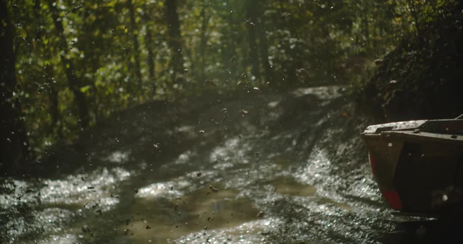 Two people ride an atv through muddy terrain in the woods during a midday sports event kicking up dirt as they navigate the rugged path surrounded by trees