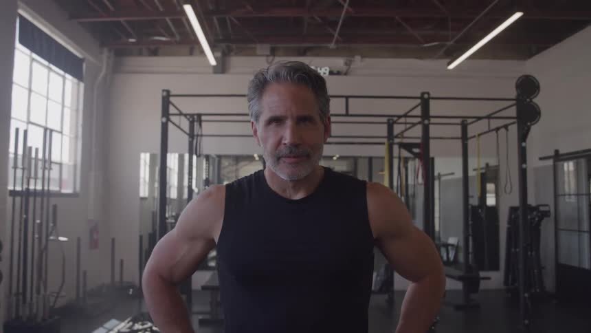 A middleaged man with gray hair and a short beard stands in a gym wearing a black tank top and a dark wristwatch he looks directly at the camera with a determined steady expression