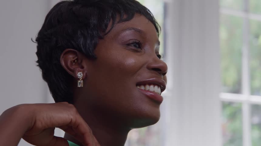 A woman with short hair smiles warmly in profile resting her hand near her chin and wearing sparkling earrings