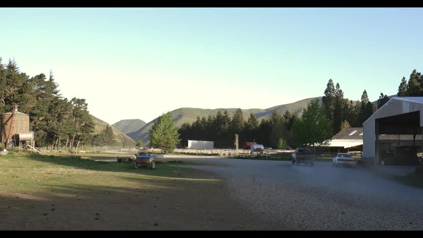 Pickup truck kicks up dust while crossing farm with silos fences and forested hills in the background