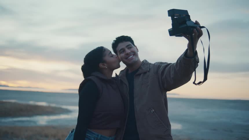 A smiling young man and a young woman pose for a selfportrait video while outdoors with the woman leaning in to kiss his cheek as he holds a vintage instant camera up to take a picture against a blurred background of a large lake and a pale sunset sky