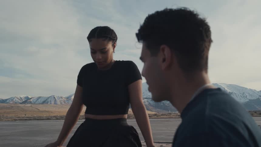 A young woman stands by the shore with snowy mountains behind gazing down with a serious thoughtful look while a young man in profile looks down quietly