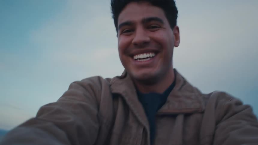 A young man with dark hair wearing a brown jacket smiles broadly into the camera in a firstperson style video with a pale blue and cloudy sky moving rapidly in the background
