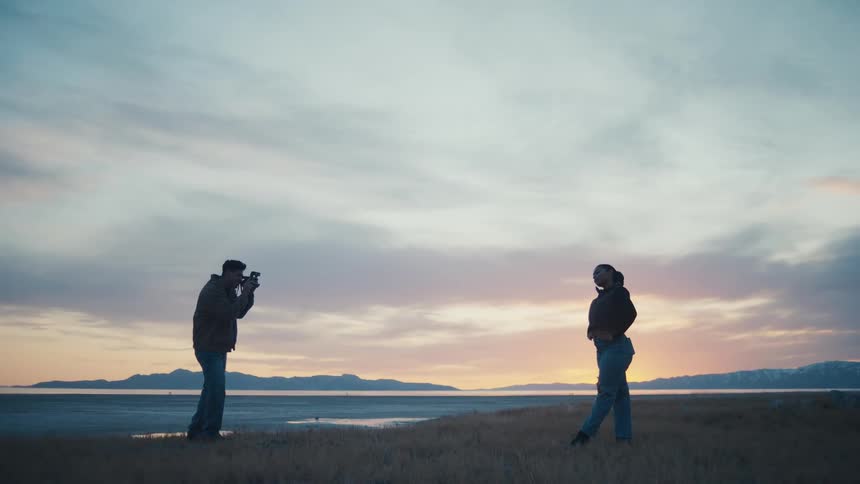At dusk by a quiet lakeshore a man with a camera photographs a woman posing playfully against a pastel sky and distant mountains