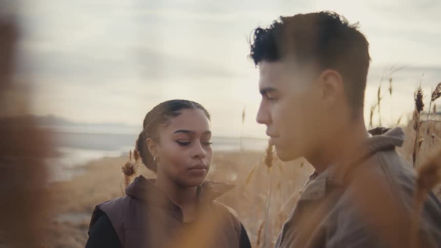 A young man and a young woman look intently at each other framed closely in a field of tall dry reeds by a body of water with bright sunlight coming from behind them giving the moment a serious and intimate atmosphere