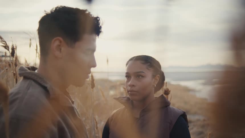 A young couple stands on a grassy shoreline at midday both wearing jackets and having a discussion