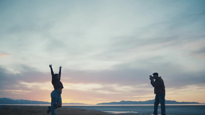 A joyful woman leaps with arms raised at sunset while a man stands a short distance away focused on photographing her against a wide lakeside and mountain backdrop