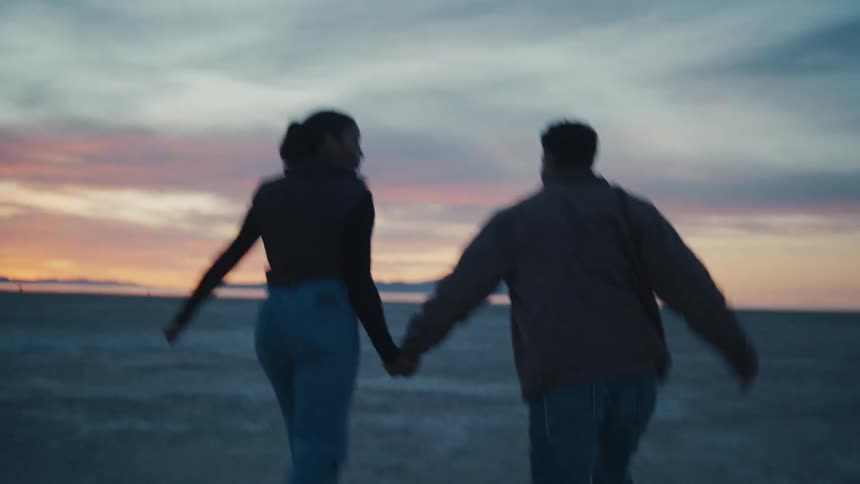 A young couple run away from the camera holding hands and smiling in silhouette against a pale blue and orange sunset sky over a vast flat salt desert or dried lake bed