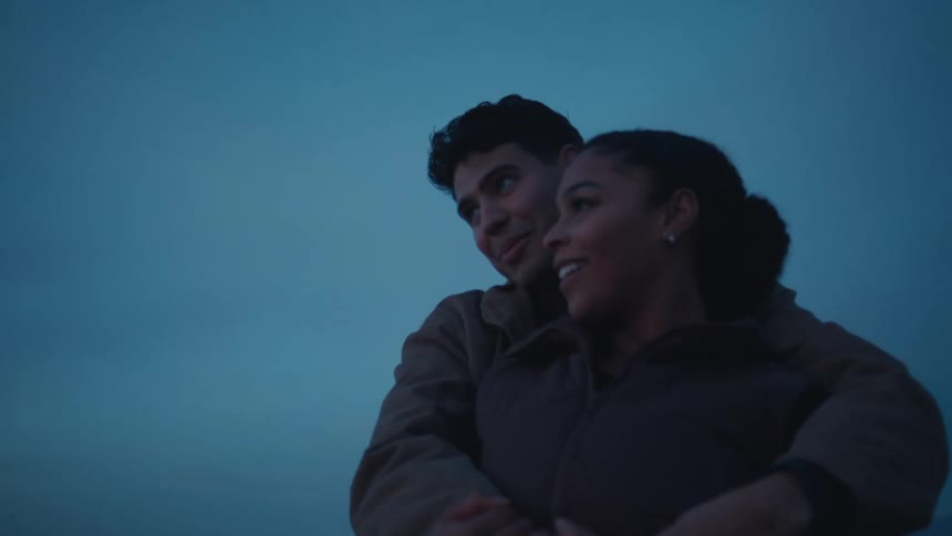 At dusk a man embraces a woman from behind as they look out together their expressions soft and affectionate
