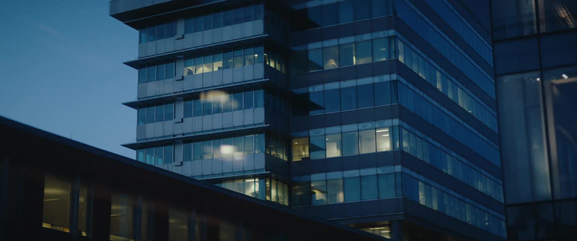 A modern glass office tower at dusk glows with scattered interior lights its stacked corners and reflections giving a cool corporate skyline feel