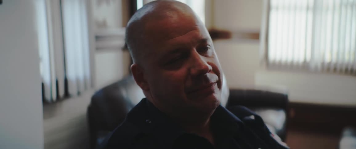 A medium closeup shot shows a middleaged bald man with a serious expression likely wearing a uniform or dark collared shirt he is seated indoors looking slightly offcamera to the right with vertical blinds visible in the background