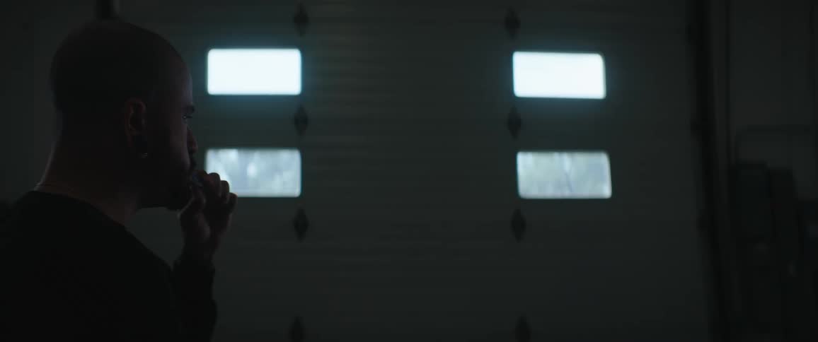 A man stands in a dim garage facing a closed door with four glowing windows silhouetted as he brushes his teeth