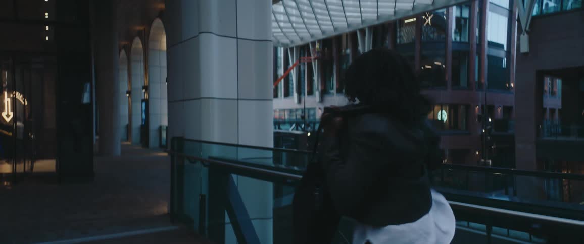 A person with curly hair walks along an elevated glass walkway inside a modern atrium at duskwhile putting a jacket on