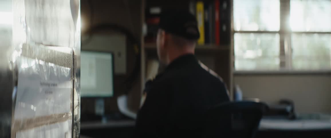 Man in uniform wearing a baseball cap sitting on a chair working on a computer in an office indoors during day time