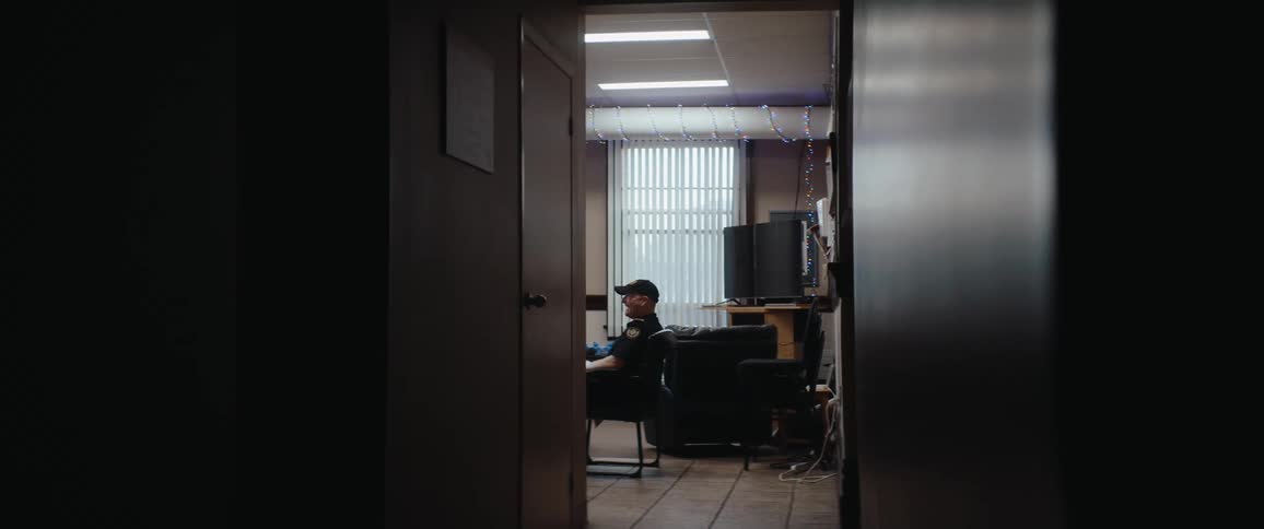 The video is an interior shot framed by a dark doorway looking into a dimly lit office or lounge area where a person in a black cap and dark shirt is seateda string of what appear to be purple fairy lights is visible across the top of the wal