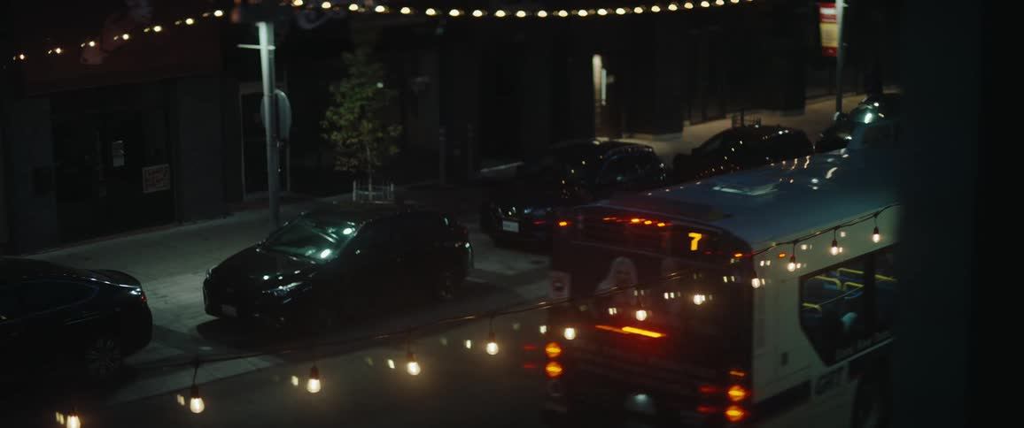 A series of cars are parked along the side of the street at night while other cars drive past under the streetlights