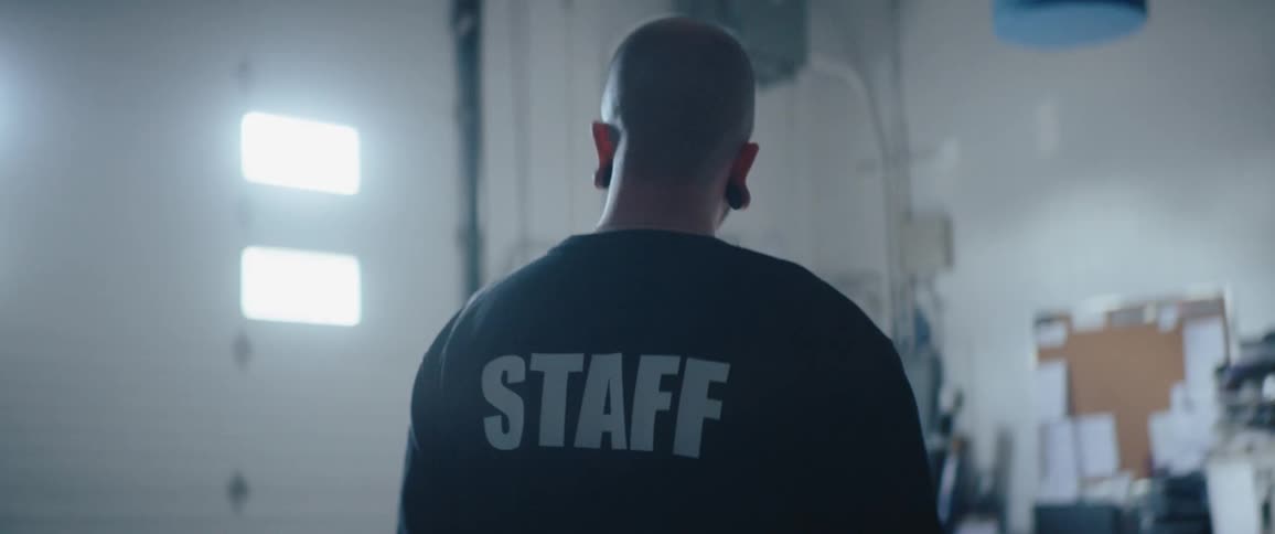 The video is an overtheshoulder shot from behind a person wearing a black shirt with the word staff printed in white across the upper backthe person is bald and standing in an industrial or gymlike setting turning to the right to look offcam