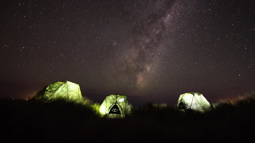The provided video clip shows three illuminated tents set up in a dark grassy field above the campsite the milky way and a dense field of stars are visible in the night sky