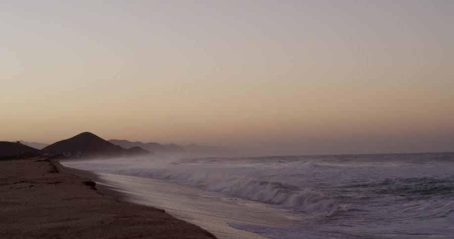 The video is a wide long shot of a beach at what appears to be sunrise or sunset with mountains visible in the distance on the left waves are crashing onto the shore along the right side of the frame