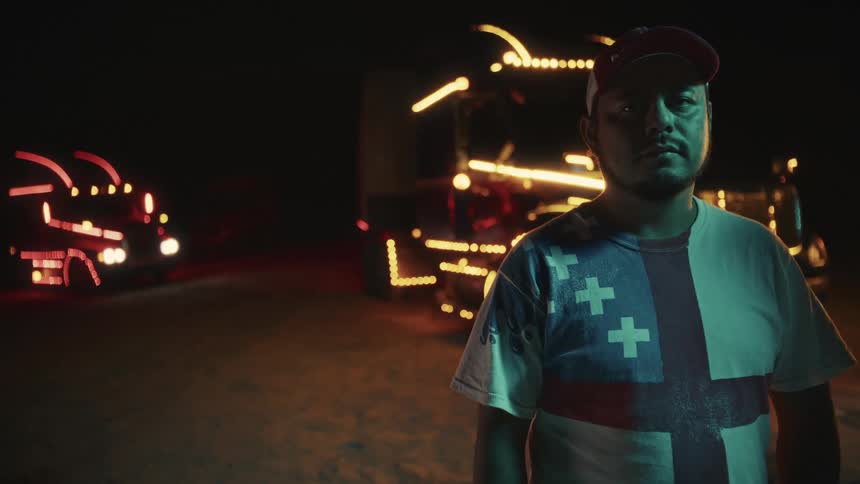 The video shows a man standing outdoors at night looking directly at the camera with a neutral expression he is wearing a cap and a tshirt featuring a distinctive design of red white and blue with three white crosses which resembles the flag of amst