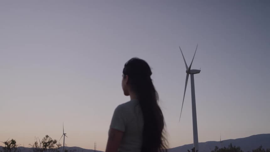 At dusk a woman gazes thoughtfully toward a towering wind turbine on the horizon her posture calm and contemplative