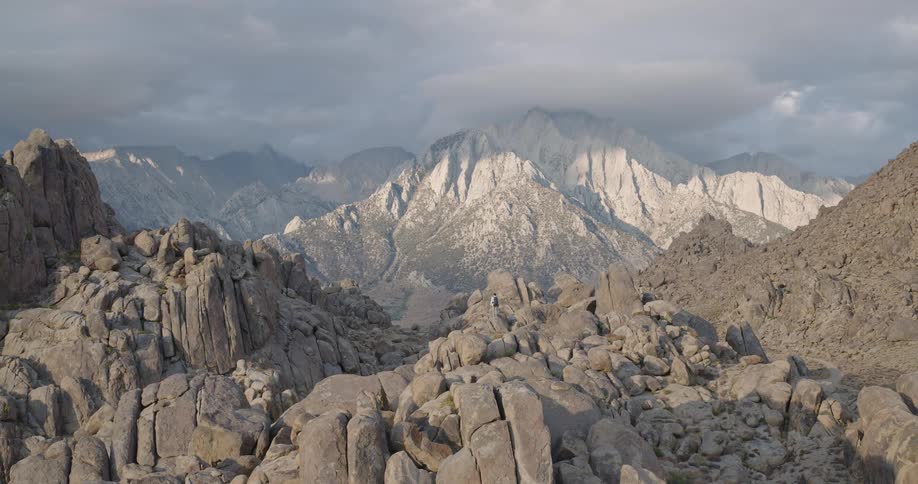 An aerial video slowly reveals a male hiker standing on a rocky outcrop in the middle of a vast desolate field of gray boulders overlooking a deep valley toward a huge pale granite mountain range with distant snow patches under a dramatic cloudy sky