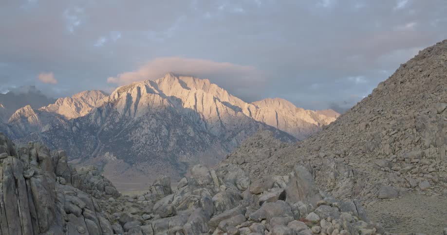 The video is a sweeping shot of a dramatic mountain range against a pale sky with the slopes bathed in golden evening sunlightthe foreground is filled with a chaotic terrain of rough light gray boulders that frame the towering peaks