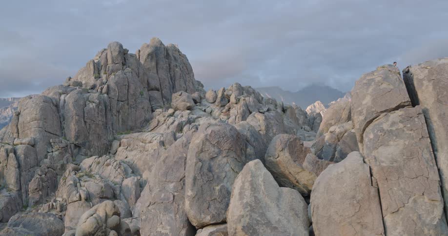 A lone hiker stands atop massive weathered boulders against a hazy mountain backdrop appearing small but purposeful