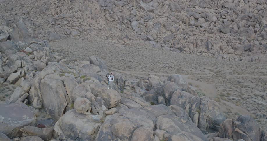 The video features a lone hiker with a backpack standing on a high point amidst a chaotic landscape of light gray cracked bouldersthe camera slowly zooms out revealing the vast scale of the rugged terrain and the imposing distant mountain