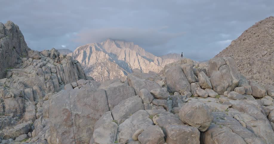 A lone hiker with a backpack navigates a vast rocky mountain landscape under a cloudy sky