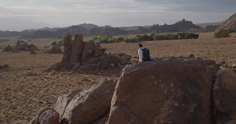 Man sitting down on a tall rock looking through binoculars at a natural landscape on a mountainous region outdoors during day time