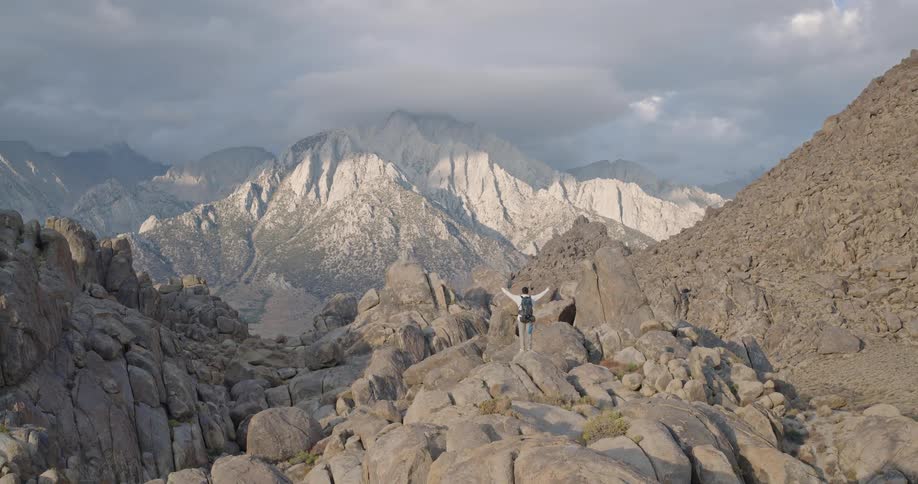 An aerial view video shows a male hiker raising his arms in triumph while standing on a large gray boulder amidst a massive rocky landscape with a distant majestic granite mountain range and cloudy sky filling the background