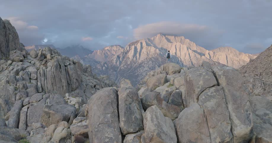 A person stands atop a rock formation in the foreground facing a distant mountain peak under a cloudy sky